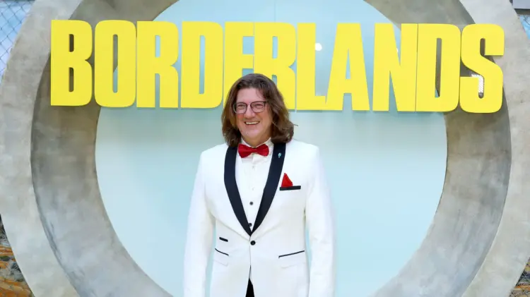 Randy Pitchford mengenakan jas putih tersenyum di depan logo Borderlands, merefleksikan asal usul bocoran aneh Pixel Watch 5.