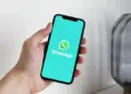 Tampilan layar ponsel cerdas yang menampilkan logo WhatsApp di atas latar belakang teal cerah, digenggam oleh tangan, mengilustrasikan daftar perangkat yang tidak lagi didukung.