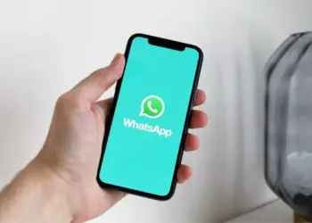 Tampilan layar ponsel cerdas yang menampilkan logo WhatsApp di atas latar belakang teal cerah, digenggam oleh tangan, mengilustrasikan daftar perangkat yang tidak lagi didukung.