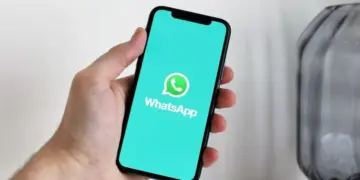 Tampilan layar ponsel cerdas yang menampilkan logo WhatsApp di atas latar belakang teal cerah, digenggam oleh tangan, mengilustrasikan daftar perangkat yang tidak lagi didukung.