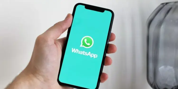 Tampilan layar ponsel cerdas yang menampilkan logo WhatsApp di atas latar belakang teal cerah, digenggam oleh tangan, mengilustrasikan daftar perangkat yang tidak lagi didukung.