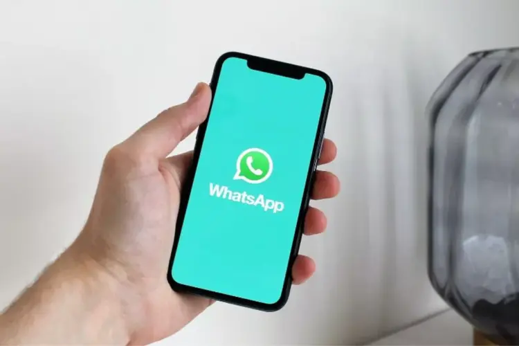 Tampilan layar ponsel cerdas yang menampilkan logo WhatsApp di atas latar belakang teal cerah, digenggam oleh tangan, mengilustrasikan daftar perangkat yang tidak lagi didukung.
