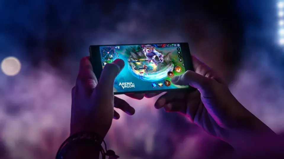 Kedua tangan memegang smartphone dalam posisi lanskap, menampilkan gameplay Arena of Valor dengan pencahayaan neon estetik.