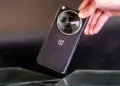 Sebuah tangan memegang smartphone OnePlus hitam dengan tekstur bodi bergaris halus dan modul kamera melingkar berlogo H.