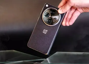 Sebuah tangan memegang smartphone OnePlus hitam dengan tekstur bodi bergaris halus dan modul kamera melingkar berlogo H.
