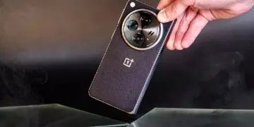 Sebuah tangan memegang smartphone OnePlus hitam dengan tekstur bodi bergaris halus dan modul kamera melingkar berlogo H.