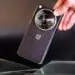 Sebuah tangan memegang smartphone OnePlus hitam dengan tekstur bodi bergaris halus dan modul kamera melingkar berlogo H.