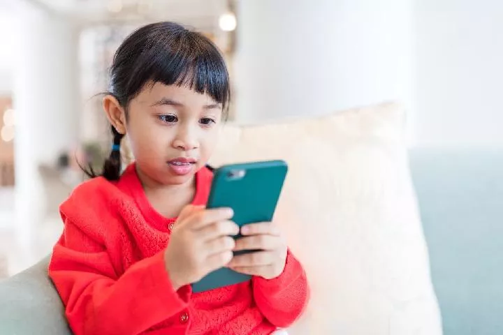 Seorang anak perempuan berambut kuncir dua mengenakan baju merah sedang fokus melihat layar HP dengan casing hijau di atas sofa.