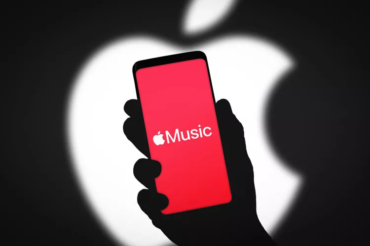Siluet tangan memegang smartphone berlayar merah dengan logo Apple Music dan latar belakang logo Apple besar.