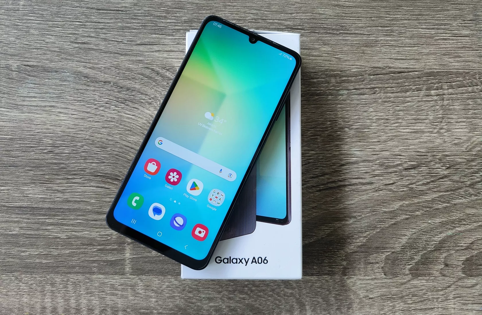 Smartphone Samsung Galaxy A06 dengan layar menyala diletakkan di atas kotak penjualan resminya di atas meja kayu.