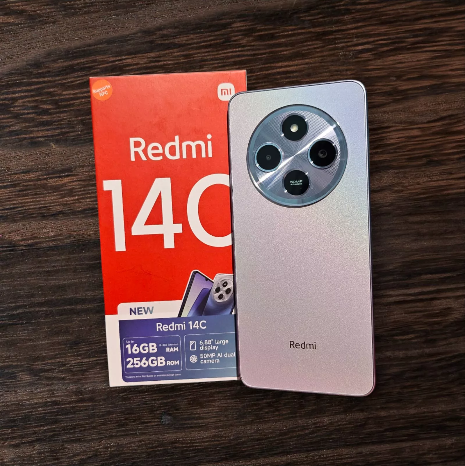 Unit Redmi 14C di atas meja kayu berdampingan dengan dus kemasan merah bertuliskan Redmi 14C.