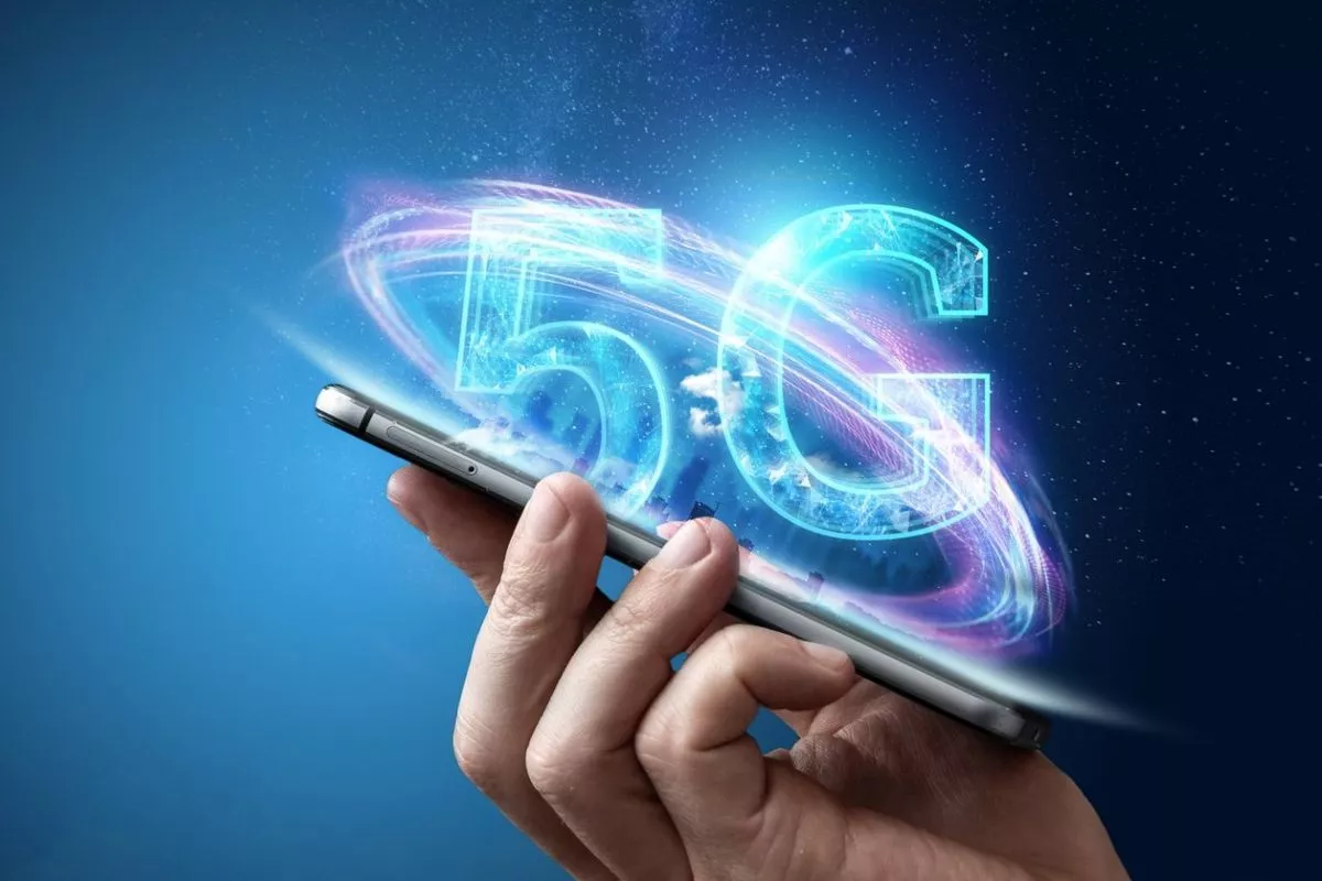 Ilustrasi tangan memegang smartphone 5G terlaris dengan efek cahaya logo 5G yang futuristik.