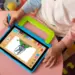 Seorang anak sedang menggunakan stylus pen untuk menggambar di tablet anak murah berukuran 2 jutaan yang dilengkapi casing pelindung tebal berwarna biru-hijau dengan pegangan tangan.
