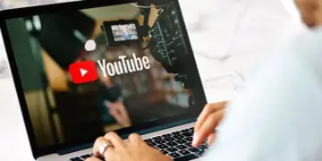 Seseorang sedang mengetik di laptop yang menampilkan logo YouTube dan peralatan studio video.