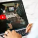 Seseorang sedang mengetik di laptop yang menampilkan logo YouTube dan peralatan studio video.