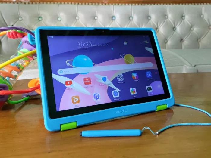 Huawei MatePad Kids Edition dengan casing pelindung biru terintegrasi, penyangga hijau, dan stylus pen khusus anak di atas meja kayu.