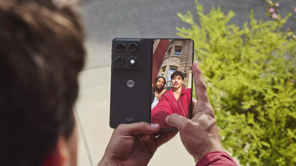 Seseorang dari sudut pandang belakang sedang menggenggam Motorola Razr Fold dalam kondisi terlipat untuk mengambil foto selfie dua pria menggunakan layar sekunder luar.