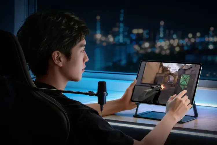 Seorang gamer pria sedang bermain game FPS (First-Person Shooter) menggunakan tablet iQOO yang diletakkan di atas stand meja saat malam hari.