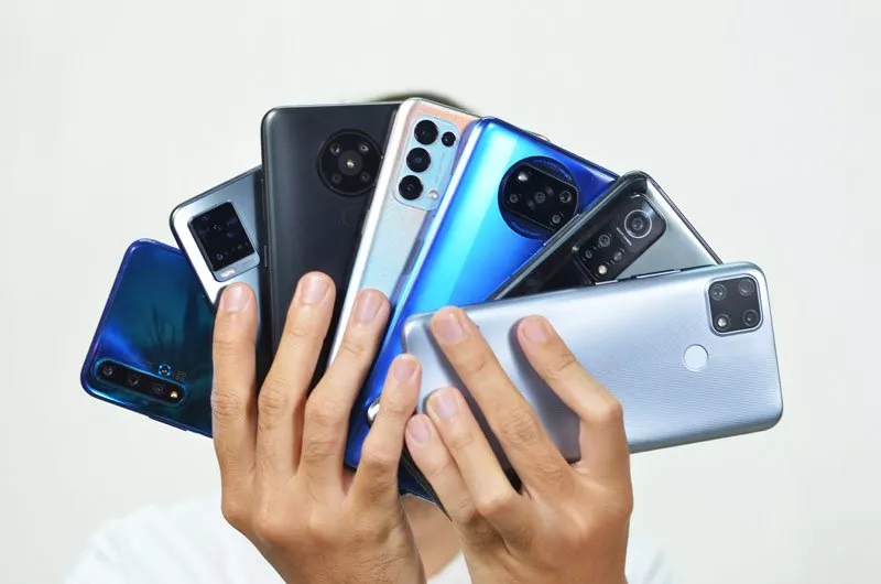 Dua tangan pria memegang dan memamerkan sekaligus tujuh buah smartphone dengan berbagai variasi model kamera belakang serta warna bodi yang berbeda.