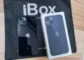Produk fisik iPhone warna hitam pekat bersanding dengan kotak retail di atas tas belanja iBox resmi