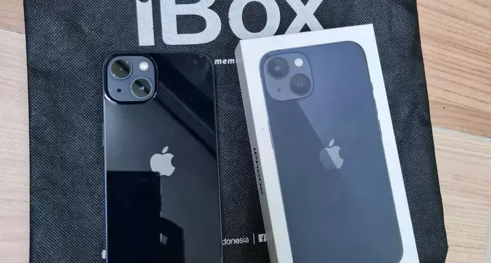 Produk fisik iPhone warna hitam pekat bersanding dengan kotak retail di atas tas belanja iBox resmi