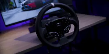 Roda kemudi sim racing dari paket Moza R5 Bundle yang terpasang di meja dengan latar belakang monitor komputer.