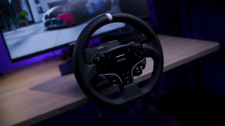 Roda kemudi sim racing dari paket Moza R5 Bundle yang terpasang di meja dengan latar belakang monitor komputer.