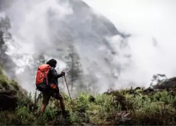 Pendaki gunung memegang trekking pole berjalan menembus kabut, membawa perlengkapan keselamatan termasuk Nitecore NWE10.