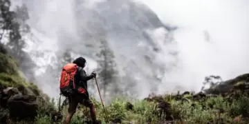 Pendaki gunung memegang trekking pole berjalan menembus kabut, membawa perlengkapan keselamatan termasuk Nitecore NWE10.