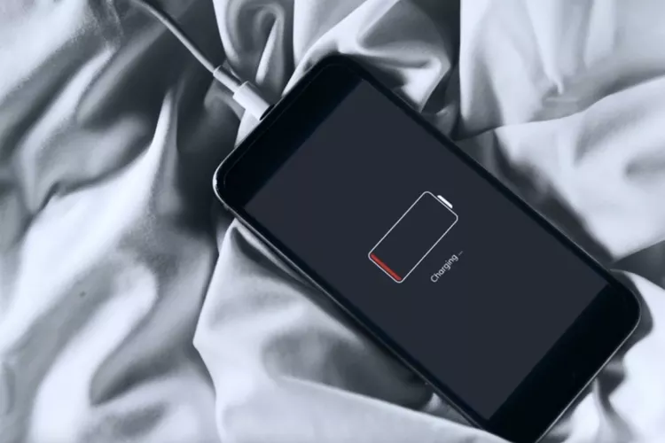 iPhone tergeletak di atas kain putih sedang diisi daya dengan indikator baterai lemah berwarna merah di layar.