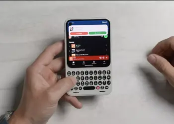 Dua tangan sedang mengoperasikan HP Android putih ber-keyboard QWERTY fisik di atas latar meja putih.