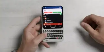 Dua tangan sedang mengoperasikan HP Android putih ber-keyboard QWERTY fisik di atas latar meja putih.