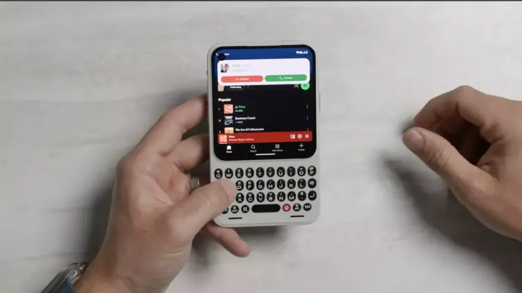Dua tangan sedang mengoperasikan HP Android putih ber-keyboard QWERTY fisik di atas latar meja putih.