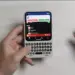 Dua tangan sedang mengoperasikan HP Android putih ber-keyboard QWERTY fisik di atas latar meja putih.
