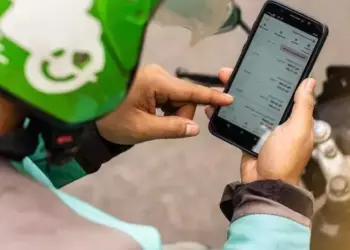Tangan driver ojol mengenakan helm hijau sedang menekan layar smartphone yang menampilkan daftar aplikasi HP untuk Ojol.