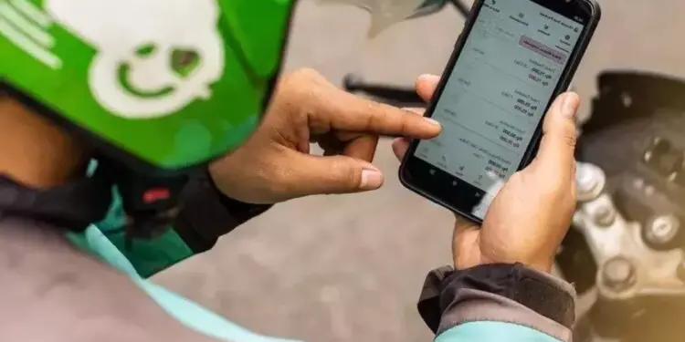 Tangan driver ojol mengenakan helm hijau sedang menekan layar smartphone yang menampilkan daftar aplikasi HP untuk Ojol.