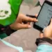 Tangan driver ojol mengenakan helm hijau sedang menekan layar smartphone yang menampilkan daftar aplikasi HP untuk Ojol.