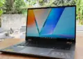 Laptop ASUS Vivobook 14 Flip TP3407AA terbuka di atas meja kayu dengan layar OLED berwarna cerah.