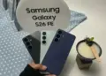 Seseorang memegang tiga varian warna Samsung Galaxy S26 FE di depan stand bertuliskan "Samsung Galaxy S26 FE".