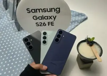 Seseorang memegang tiga varian warna Samsung Galaxy S26 FE di depan stand bertuliskan "Samsung Galaxy S26 FE".