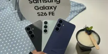 Seseorang memegang tiga varian warna Samsung Galaxy S26 FE di depan stand bertuliskan "Samsung Galaxy S26 FE".
