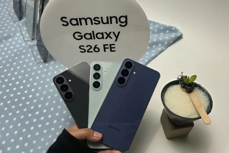 Seseorang memegang tiga varian warna Samsung Galaxy S26 FE di depan stand bertuliskan "Samsung Galaxy S26 FE".
