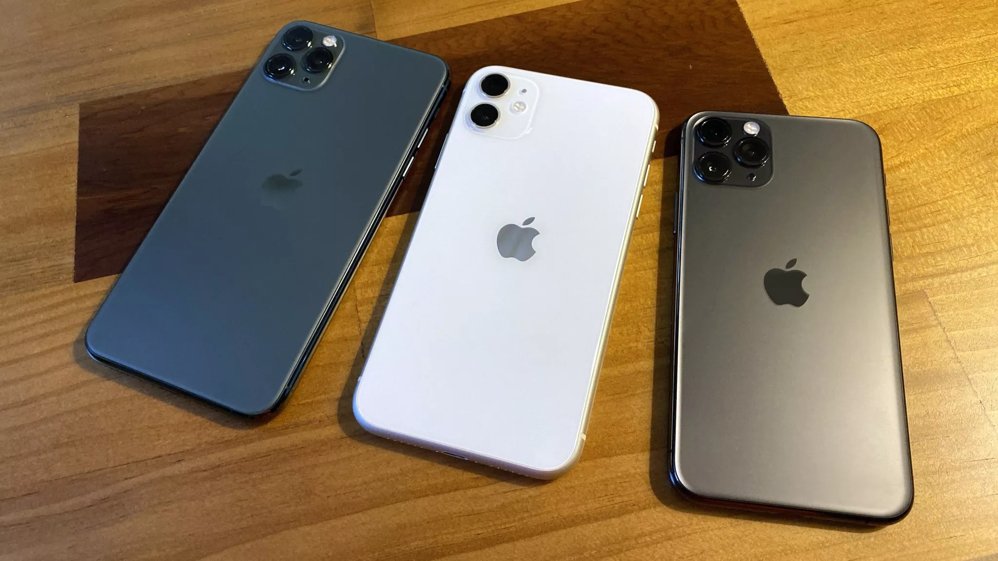 Tiga unit iPhone 11 series bekas warna Midnight Green, Putih, dan Space Gray di atas meja kayu.