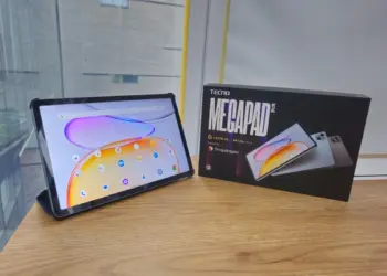 Wujud asli unit Tecno Megapad SE 2 menyala di samping kotak kemasan resminya di atas meja kayu.