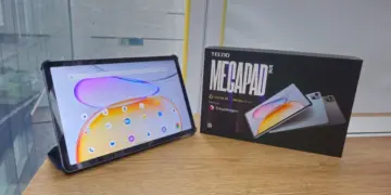 Wujud asli unit Tecno Megapad SE 2 menyala di samping kotak kemasan resminya di atas meja kayu.