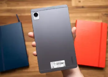 Sisi belakang tablet realme Pad Mini warna abu-abu yang digenggam satu tangan di atas meja kayu.