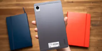 Sisi belakang tablet realme Pad Mini warna abu-abu yang digenggam satu tangan di atas meja kayu.