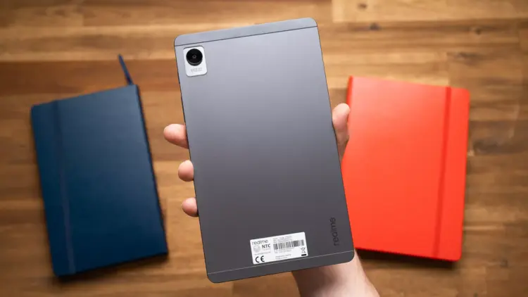 Sisi belakang tablet realme Pad Mini warna abu-abu yang digenggam satu tangan di atas meja kayu.