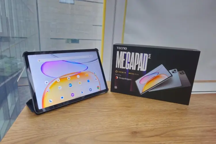 Wujud asli unit Tecno Megapad SE 2 menyala di samping kotak kemasan resminya di atas meja kayu.
