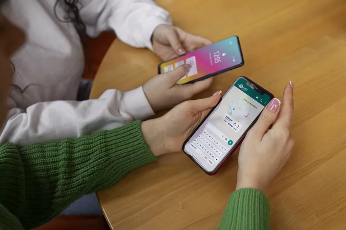 Dua orang memegang iPhone masing-masing di atas meja kayu, menggunakan aplikasi berbeda (peta dan pesan) secara bersamaan dengan satu nomor HP melalui iOS 27.